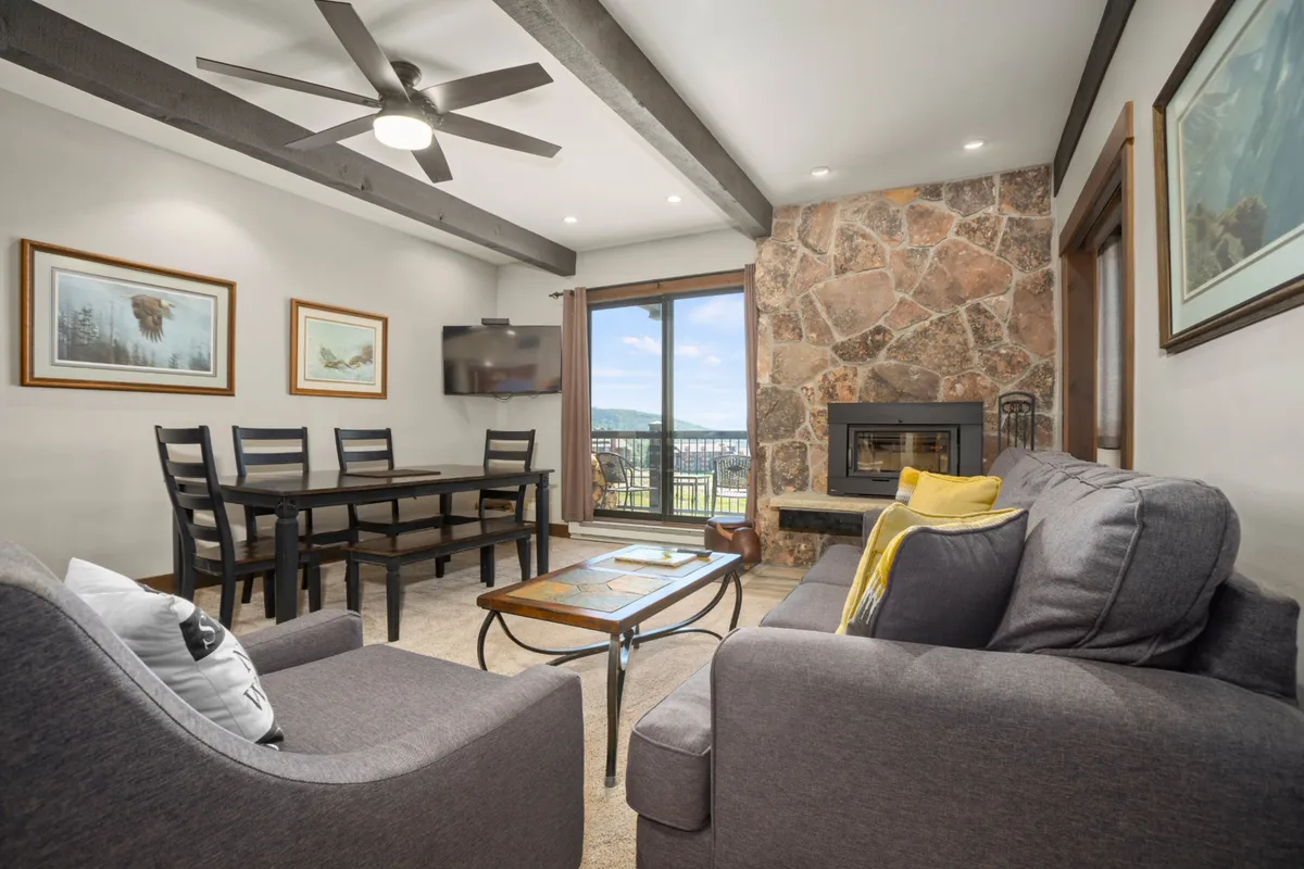 A view from the dining area shows a spacious open-plan layout with a large wooden dining table, a stone fireplace in the background, and oversized windows offering mountain vistas. The connection between dining, relaxing, and enjoying the view is visually emphasized.