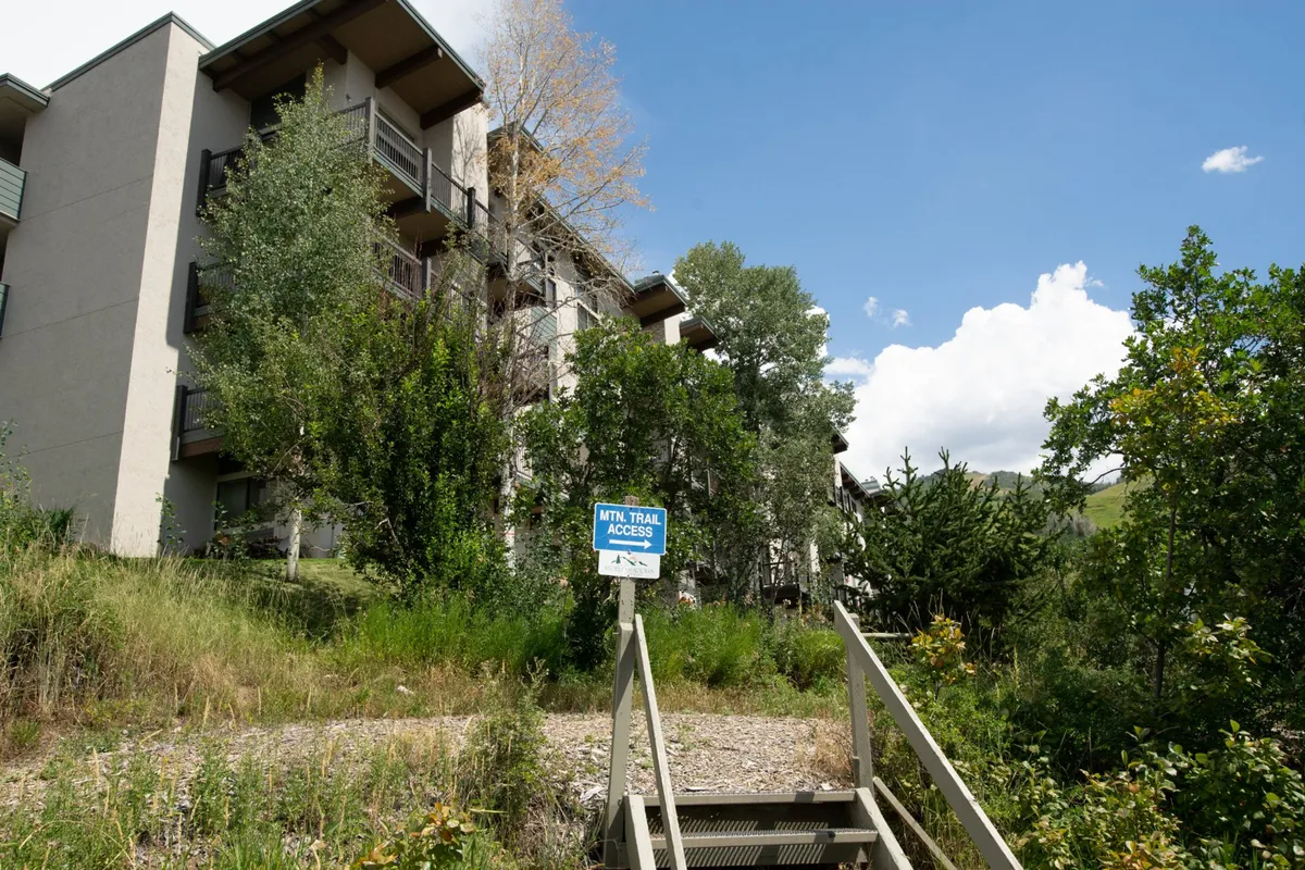 This exterior image shows the apartment building surrounded by lush greenery, with a prominent trail access sign in the foreground. The setting highlights convenient entry to hiking and nature trails, with trees and mountain scenery suggesting a tranquil, adventurous environment.