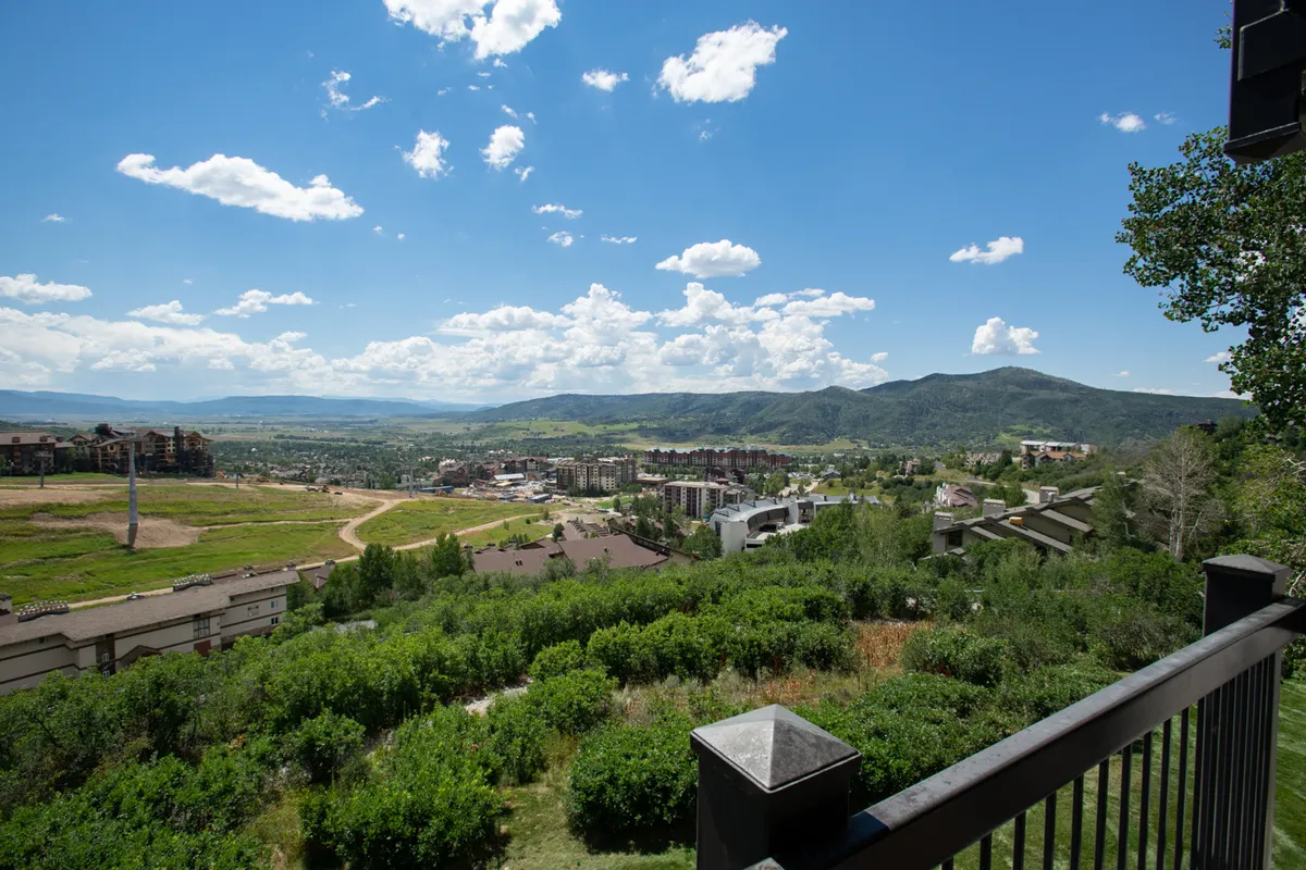 This scenic view captures a wide mountain range, the town below, lush greenery, and a dramatic sky. The vantage point could be from a balcony or a trail, evoking the beauty and tranquility of Steamboat's natural setting.