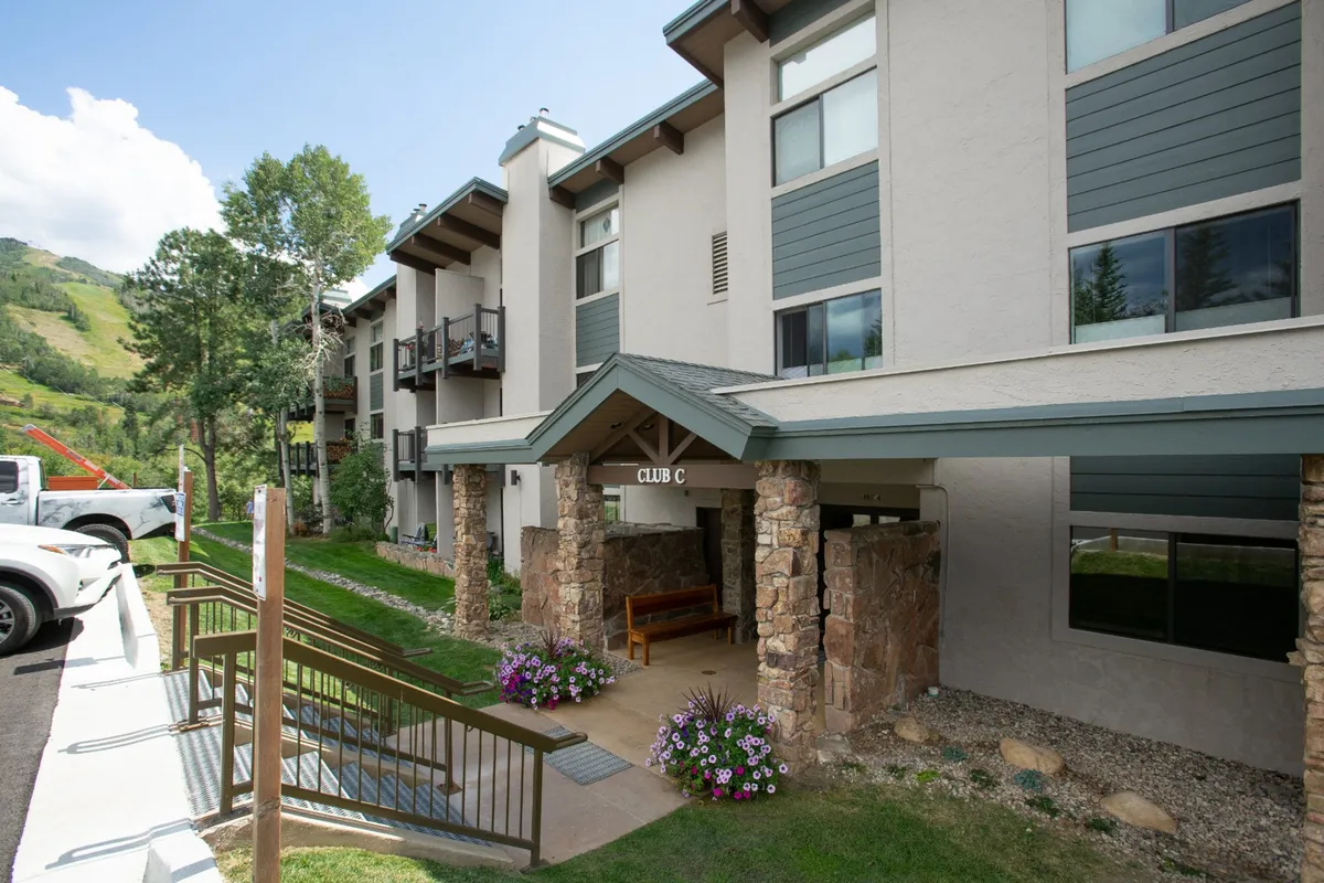 The entrance to the clubhouse, marked by a distinctive sign reading 'CLUB C' set between two stone pillars, surrounded by blooming flower beds and landscaping. The overall ambiance is inviting and hints at a well-maintained, community-oriented facility where key amenities are located.