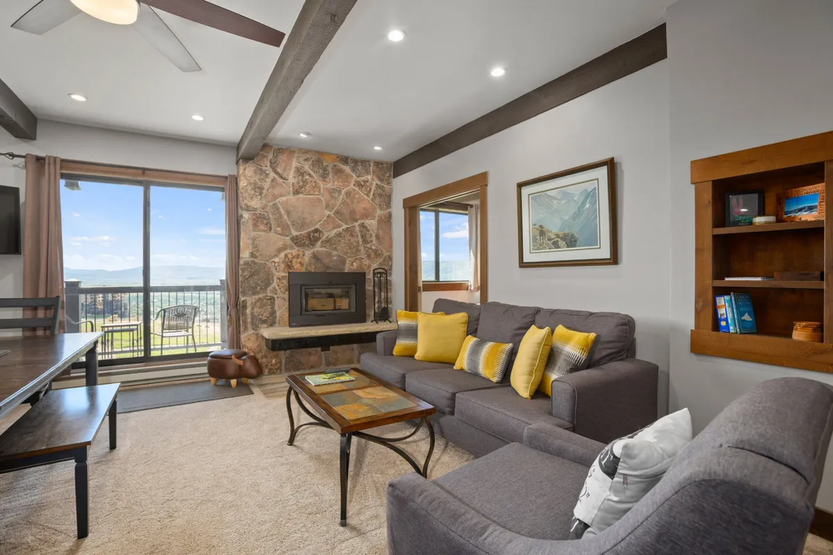 A warm and welcoming living area featuring a striking stone fireplace as the centerpiece. The space is filled with comfortable seating, decorative pillows, and large windows that offer expansive mountain views. Wooden furniture and cozy accents enhance the inviting atmosphere, making it perfect for gatherings.