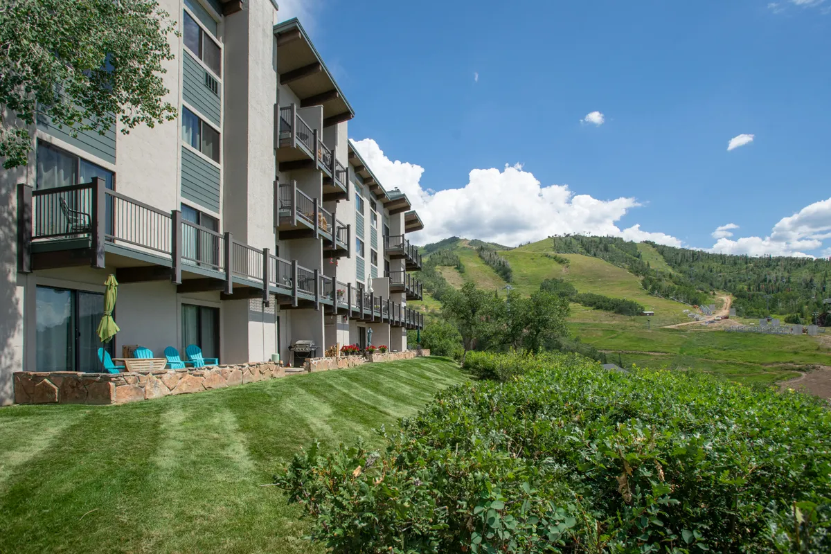 A multi-story building with balconies overlooks a manicured lawn and is framed by a dramatic mountain backdrop beneath clear blue skies. The scene emphasizes a tranquil, scenic environment and the opportunity for outdoor relaxation, with balconies inviting guests to enjoy fresh air and views.