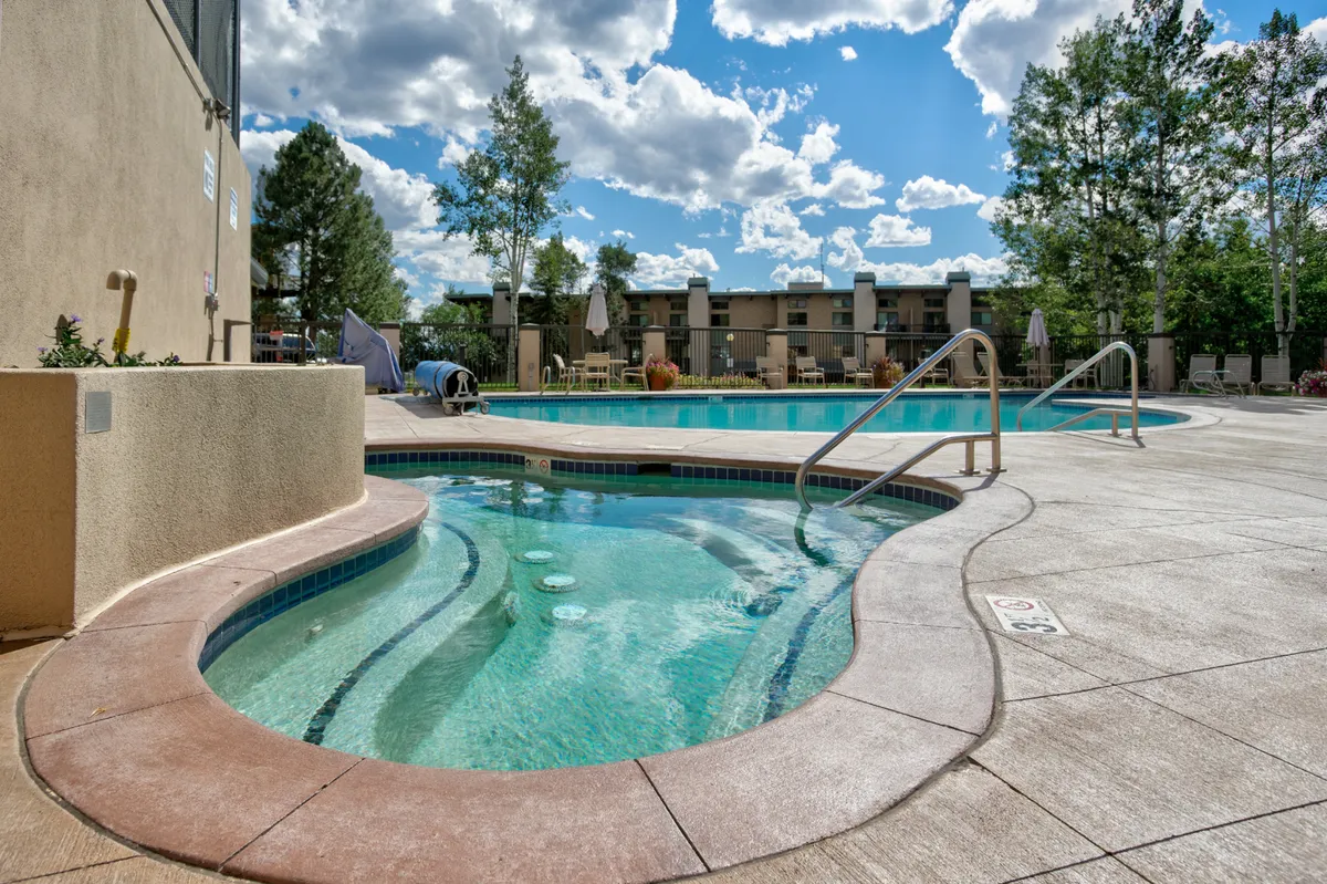 This exterior image showcases a large heated swimming pool and a bubbling hot tub surrounded by lush trees and inviting lounge chairs. The scene is lively, highlighting guests swimming, relaxing in the hot tub, sunbathing, and socializing by the pool area.
