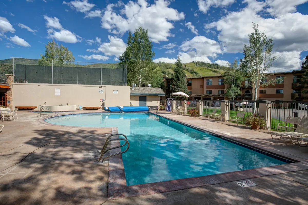 A sparkling swimming pool is surrounded by trees and lounge chairs, set against a backdrop of mountains. The image evokes a sense of relaxation and leisure, showing an inviting spot to cool off after a day of outdoor exploration.