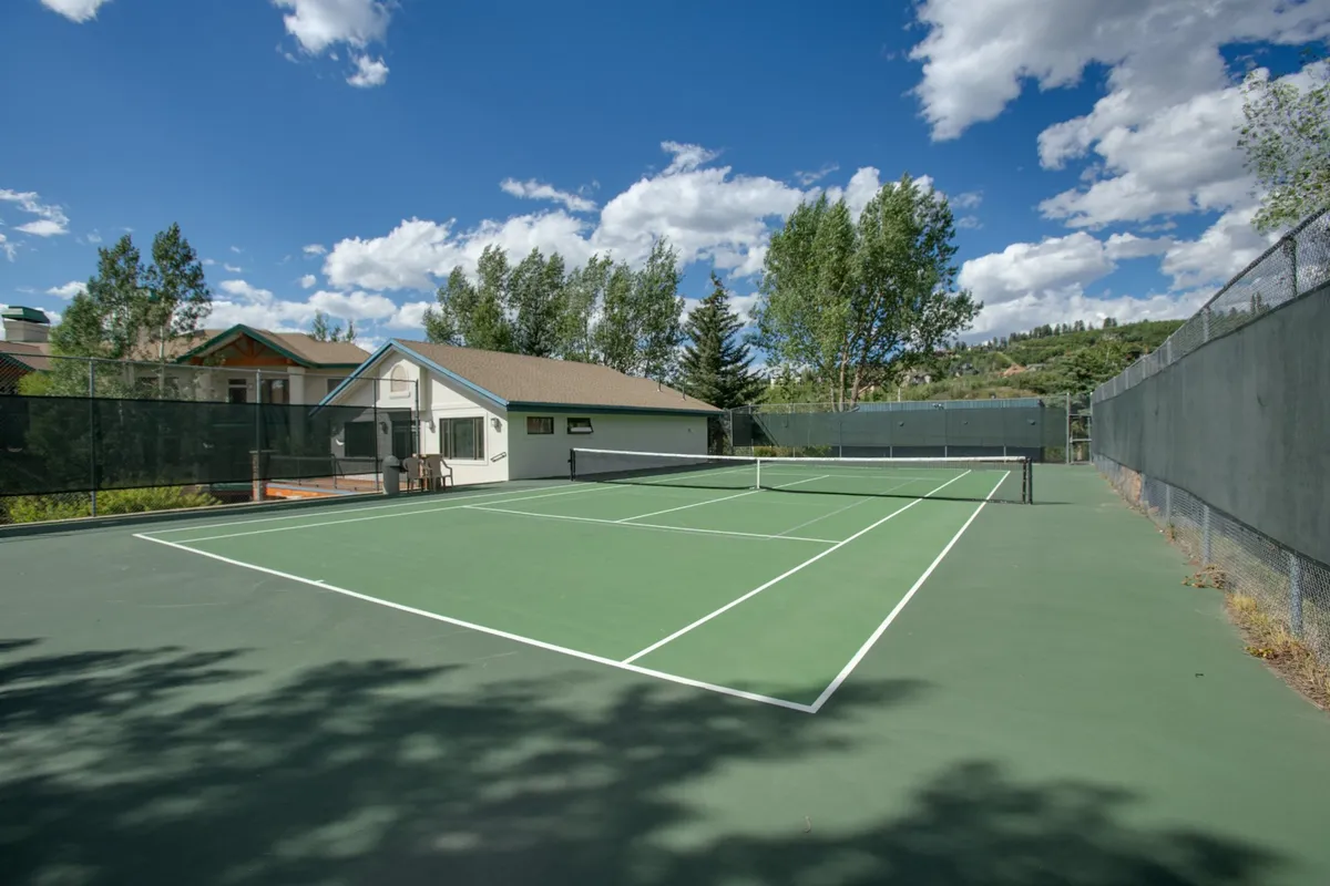 An exterior shot of a tennis court bordered by trees and nearby buildings, capturing the spirit of friendly competition and outdoor recreation. The setting hints at both social and fitness opportunities offered to guests.