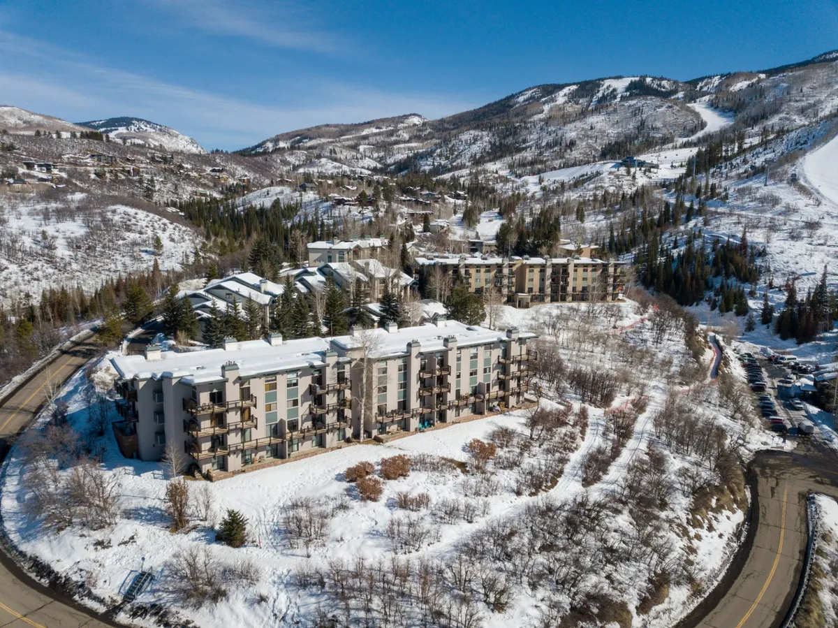 A picturesque view of snow-covered buildings nestled against a mountain backdrop with a clear blue sky. The tranquil winter landscape is accentuated by the tree line, capturing the serene and adventurous spirit of a mountain getaway.