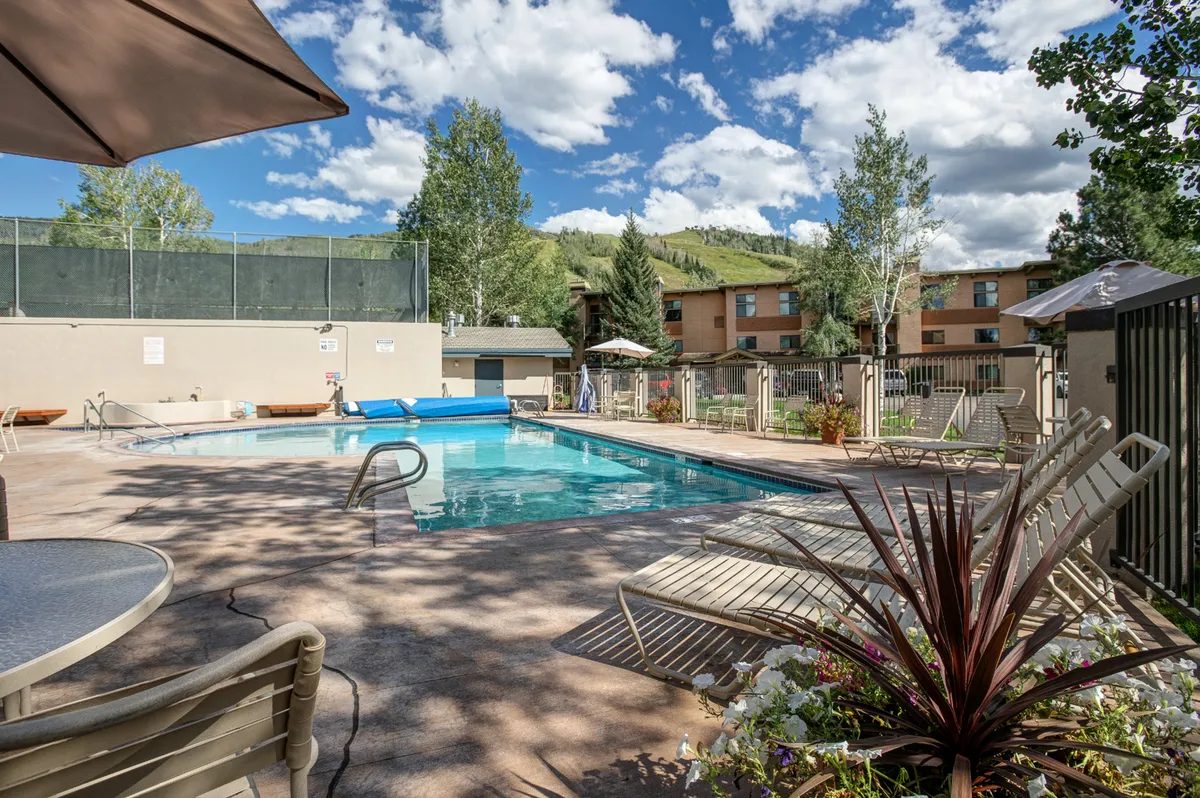 A vibrant outdoor swimming pool surrounded by lush greenery, with lounge chairs and umbrellas arranged poolside. In the background, gentle hills and the blue sky create a relaxed resort ambiance. The scene suggests a welcoming space for swimming, sunbathing, and socializing, with the pool as the central amenity.