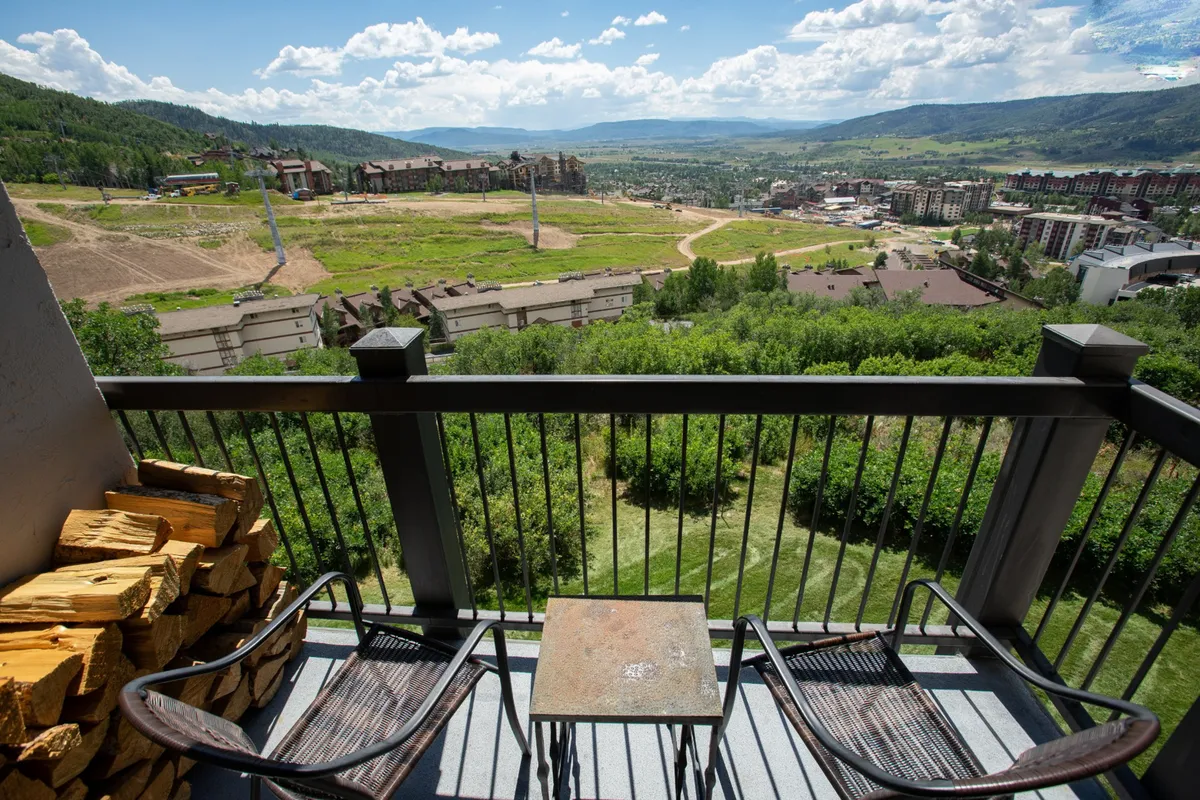 The hero image shows a private balcony overlooking a sweeping valley with majestic mountains in the distance. There are comfortable chairs and a table set for relaxation, along with a neatly stacked pile of firewood, hinting at cozy evenings by the fireplace. The scene is serene and welcoming, suggesting a perfect spot to enjoy morning coffee or watch the sun set over the peaks.