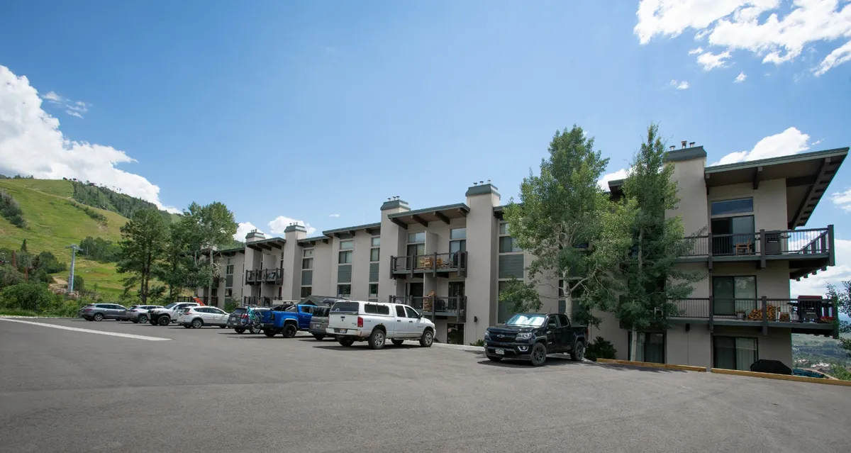 This exterior shot showcases a welcoming multi-story residential building surrounded by trees and mountains, with a visible parking area and balconies. The scene evokes a sense of arrival and community, emphasizing the ease of returning 'home' after a day spent downtown or on the slopes.