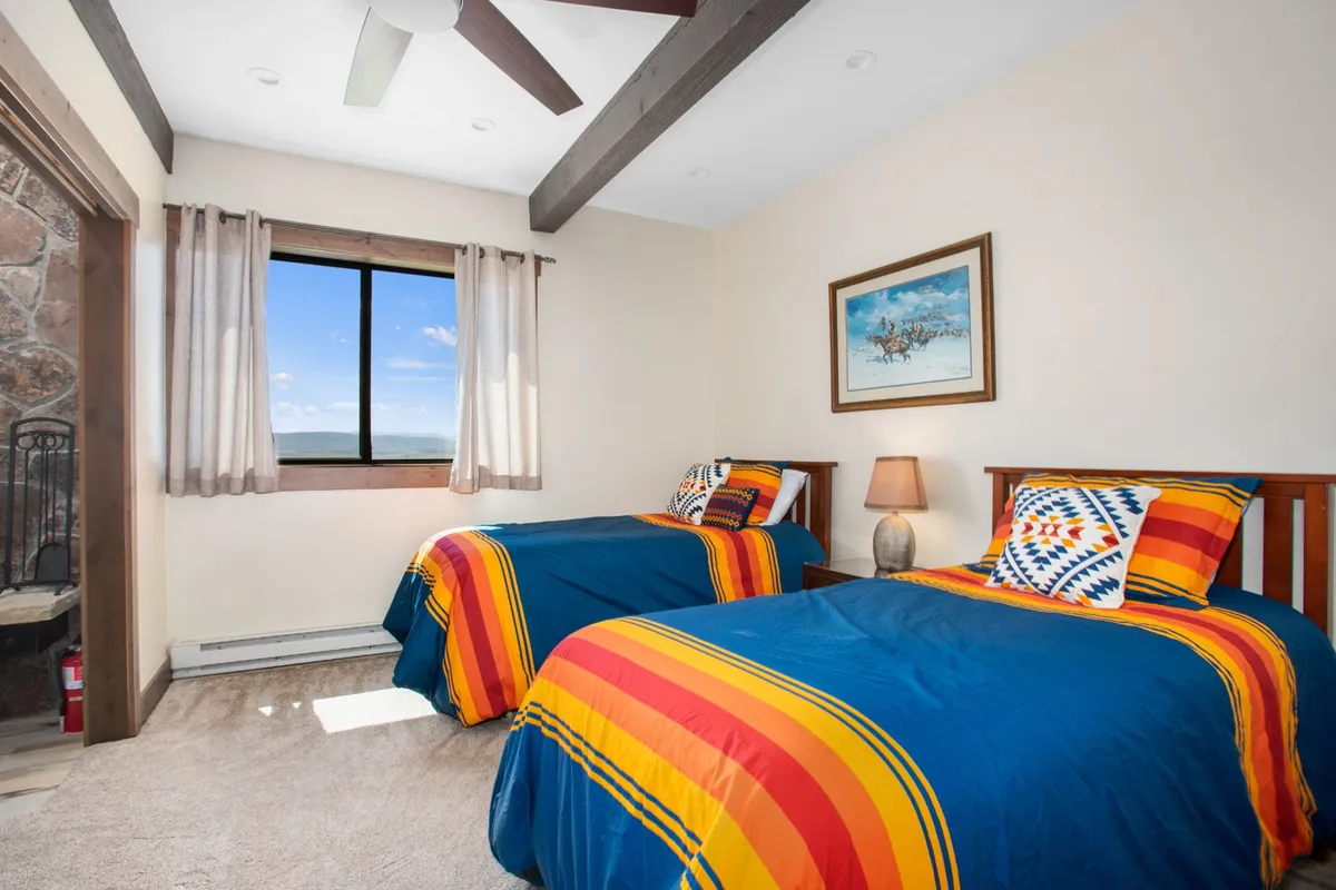 A cheerful twin bedroom featuring two beds with colorful bedding, a window that frames a scenic view, artwork on the wall, and a nightstand with a lamp. The space feels inviting and flexible for children, friends, or extra guests.