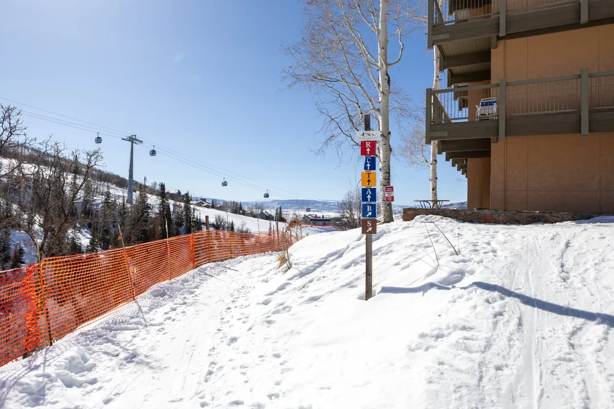 An exterior winter scene showing a snow-covered ski slope and pathway, a guidance sign indicating the ski trail, the building's exterior, and gondolas in the background. The image captures the immediate proximity to the slopes and the essence of ski-in/ski-out convenience.